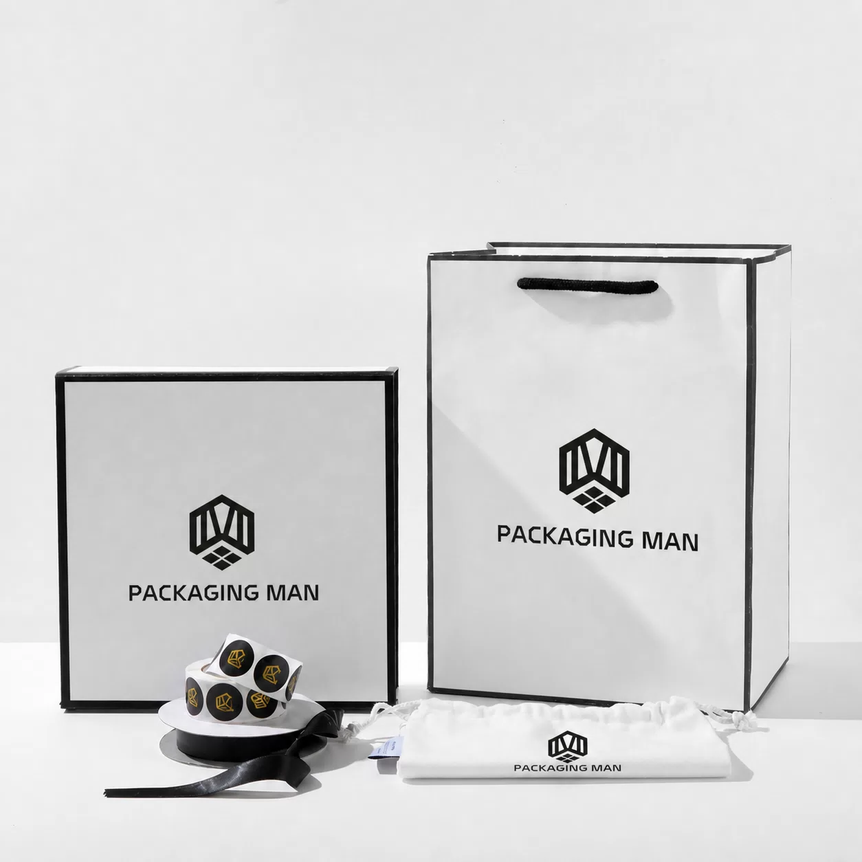 A flat lay arrangement including a square box, a tote bag, a roll of stickers, a ribbon, and a small cloth pouch.